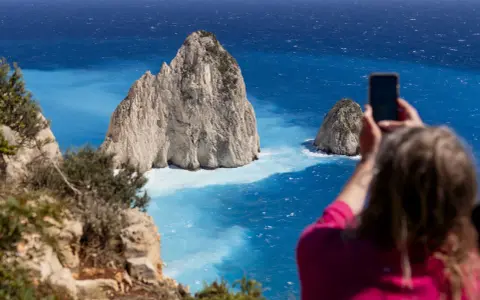 Widok na turkusowe morze i skaliste wysepki. Kobieta robi zdjęcie telefonem. Zakynthos czy warto? Tak, warto!