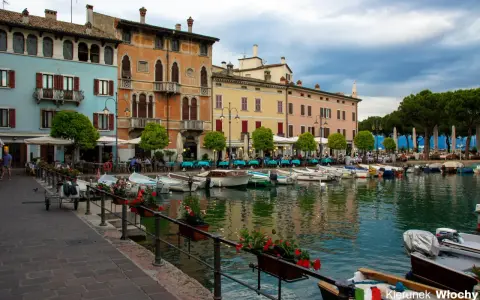 Kolorowe kamienice nad jeziorem w Desenzano del Garda, z łodziami zacumowanymi przy brzegu i stolikami restauracyjnymi na promenadzie.