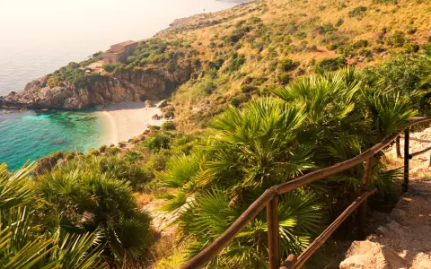 Widok na zatoczkę z piaszczystą plażą, turkusową wodą i domkiem na wzgórzu, otoczone bujną roślinnością.