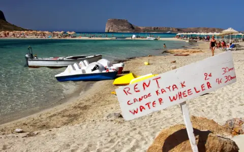 Maj na Krecie: plaża z leżakami, turkusowa woda, łódki i tablica z ofertą wynajmu kajaków i rowerów wodnych.