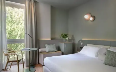 Nowoczesny pokój hotelowy z łóżkiem, fotelem i oknem z widokiem na zieleń.