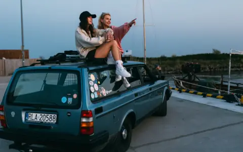 Dwie kobiety siedzą na dachu niebieskiego kombi przy nabrzeżu, jedna wskazuje palcem w dal.