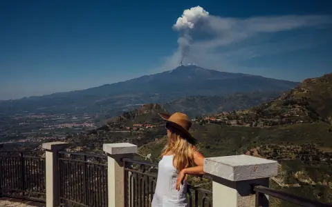Kobieta w kapeluszu patrzy na dymiący wulkan Etna z tarasu widokowego.