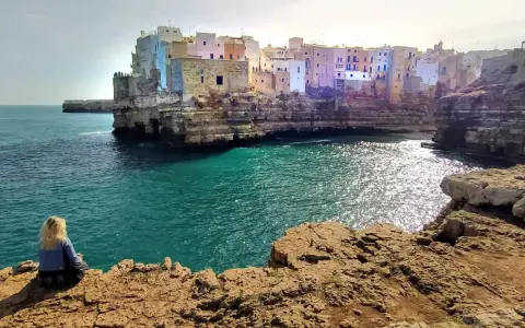 Kobieta siedzi na skale, podziwiając widok na Polignano a Mare. Białe domy wznoszą się na klifie nad turkusowym morzem.