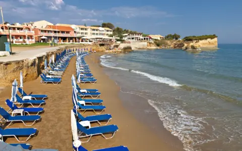 Rzędy leżaków i parasoli na piaszczystej plaży, z hotelami w tle. Idealne miejsce, by odpocząć i zastanowić się, gdzie na Korfu wybrać się na wakacje.