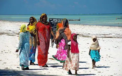 Kobiety w kolorowych strojach spacerują po plaży Zanzibaru. Warto wiedzieć, że to miejsce oferuje piękne widoki i lokalną kulturę.