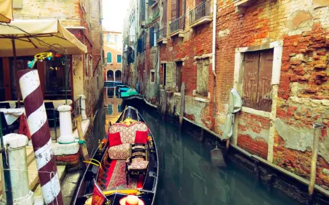 Gondola z czerwonymi poduszkami i dywanem w wąskim kanale Wenecji, otoczonym starymi ceglanymi budynkami.