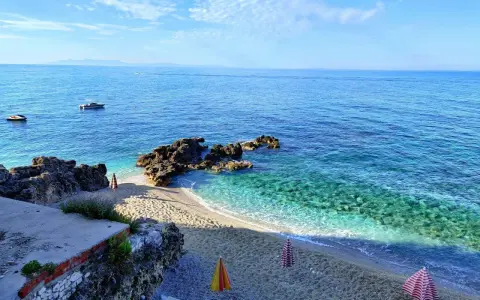 Albania, czerwiec: słoneczna pogoda nad krystalicznie czystym morzem, kamienista plaża z parasolami i łódkami na horyzoncie.