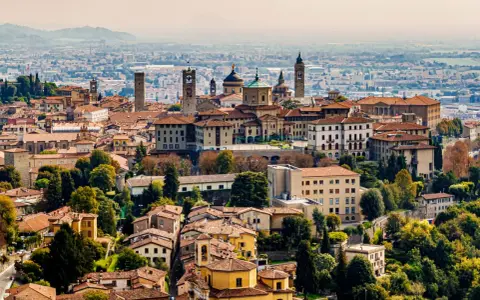 Widok na **bergamo stare miasto** z wieżami i kopułami, otoczone zielenią i innymi budynkami.