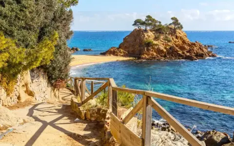 Ścieżka z drewnianą balustradą prowadzi do urokliwej zatoczki z piaszczystą plażą i skalistą wysepką. To idealne miejsce na odkrywanie uroków Costa Brava.