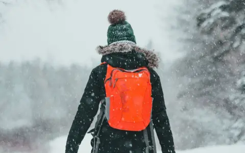 Osoba w zimowej kurtce i czapce z pomponem z pomarańczowym plecakiem idzie przez śnieg.