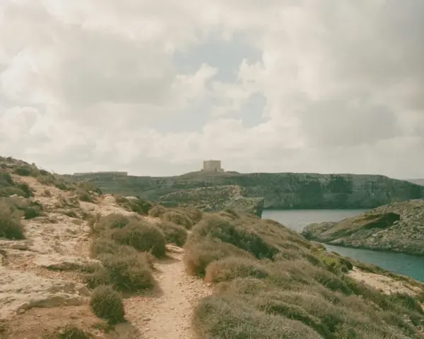 Malta czy warto? Kamienista ścieżka prowadzi przez skaliste wybrzeże z niską roślinnością do zatoki. W oddali widać wieżę obronną.