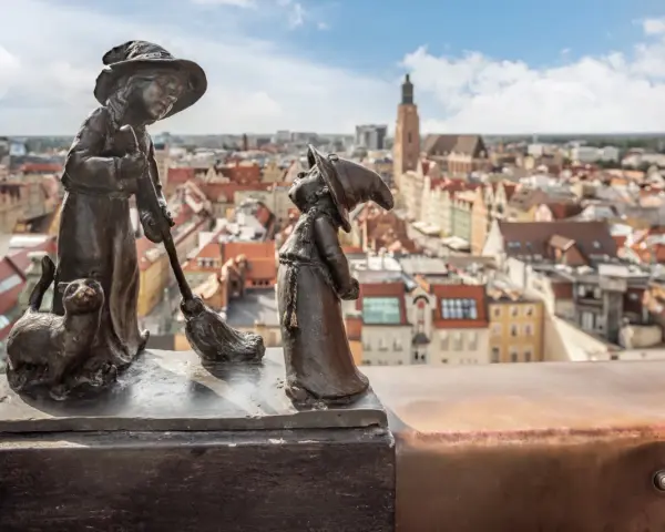Figurki krasnali: czarownica z kotem i miotłą, na tle panoramy Wrocławia.