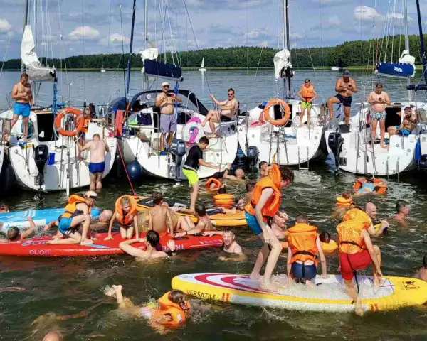 Ludzie w kamizelkach ratunkowych i strojach kąpielowych pływają i bawią się na wodzie przy żaglówkach.