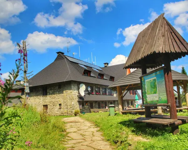 Kamienny schronisko na Turbaczu z dzieckiem w tle, otoczone zielenią i kwitnącymi kwiatami.