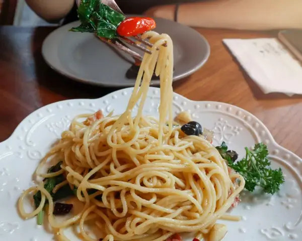 Widok z bliska na talerz z makaronem aglio olio, z widelcem unoszącym porcję spaghetti z bazylią i pomidorkiem.