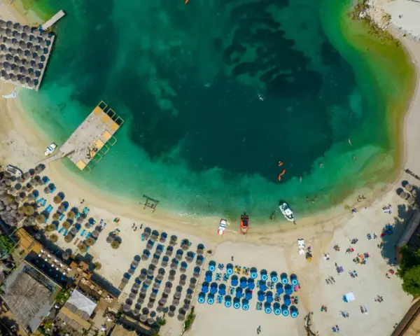 Widok z lotu ptaka na zatokę z turkusową wodą, plażą z leżakami i parasolami oraz łodziami.