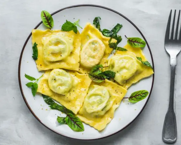 Pyszne ravioli z ricottą i szpinakiem, polane sosem, ozdobione świeżymi ziołami. Obok leży widelec.