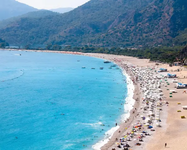 Plaża z turkusową wodą, górami w tle i tłumem ludzi odpoczywających pod parasolami.