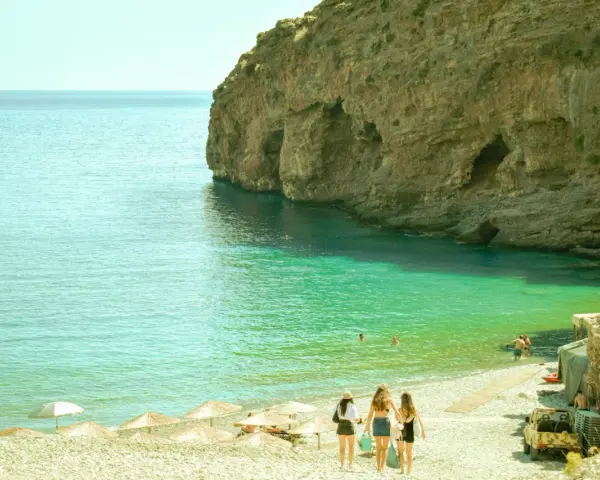 Trzy dziewczyny idące po kamienistej plaży w kierunku turkusowego morza przy skalistym klifie.
