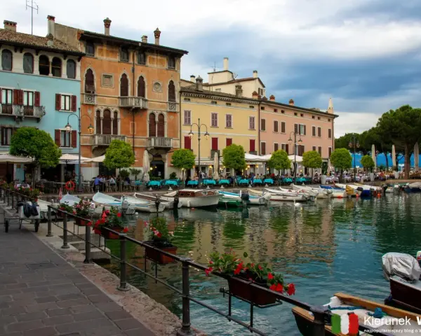 Kolorowe kamienice nad jeziorem w Desenzano del Garda, z łodziami zacumowanymi przy brzegu i stolikami restauracyjnymi na promenadzie.