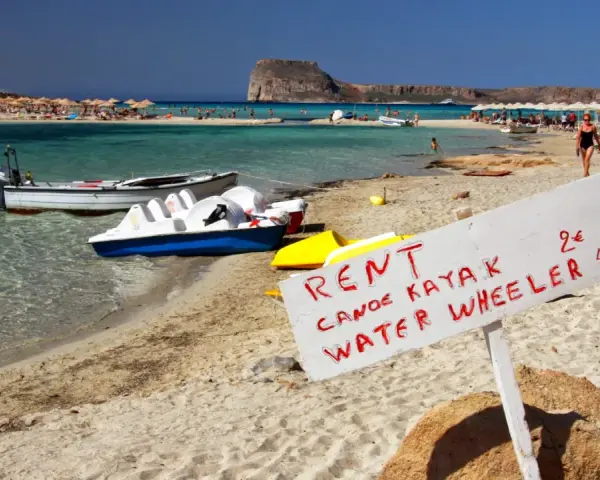 Maj na Krecie: plaża z leżakami, turkusowa woda, łódki i tablica z ofertą wynajmu kajaków i rowerów wodnych.