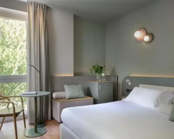 Nowoczesny pokój hotelowy z łóżkiem, fotelem i oknem z widokiem na zieleń.