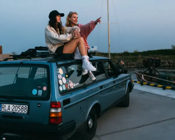 Dwie kobiety siedzą na dachu niebieskiego kombi przy nabrzeżu, jedna wskazuje palcem w dal.