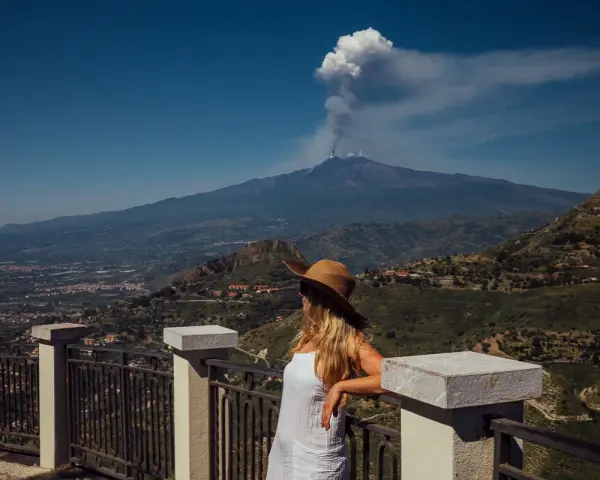 Kobieta w kapeluszu patrzy na dymiący wulkan Etna z tarasu widokowego.