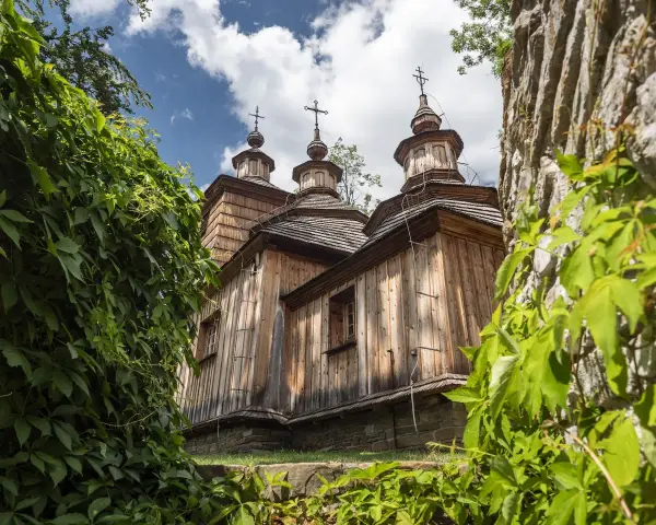 Drewniana cerkiew z trzema kopułami, otoczona zielenią, na tle błękitnego nieba z chmurami.