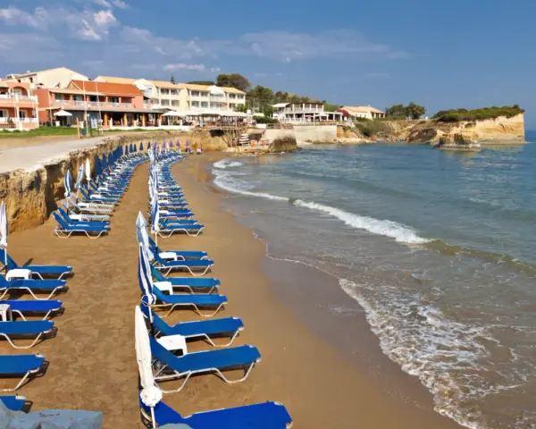 Rzędy leżaków i parasoli na piaszczystej plaży, z hotelami w tle. Idealne miejsce, by odpocząć i zastanowić się, gdzie na Korfu wybrać się na wakacje.