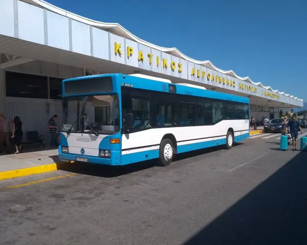 Niebiesko-biały autobus na tle budynku lotniska z żółtym napisem.