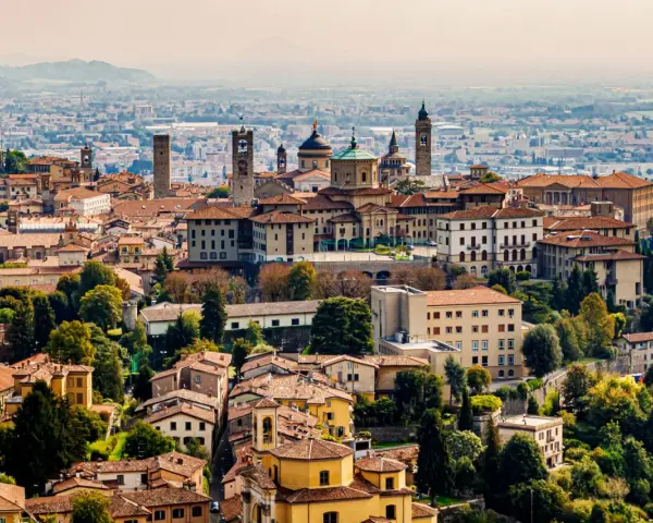 Widok na **bergamo stare miasto** z wieżami i kopułami, otoczone zielenią i innymi budynkami.