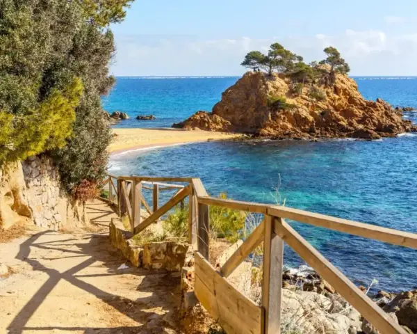 Ścieżka z drewnianą balustradą prowadzi do urokliwej zatoczki z piaszczystą plażą i skalistą wysepką. To idealne miejsce na odkrywanie uroków Costa Brava.