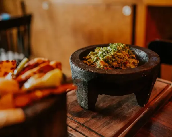 Świeże guacamole w kamiennym moździerzu, obok talerza z innymi najpopularniejszymi daniami meksykańskimi.