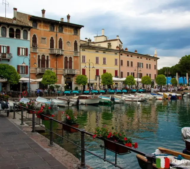 Kolorowe kamienice nad jeziorem w Desenzano del Garda, z łodziami zacumowanymi przy brzegu i stolikami restauracyjnymi na promenadzie.