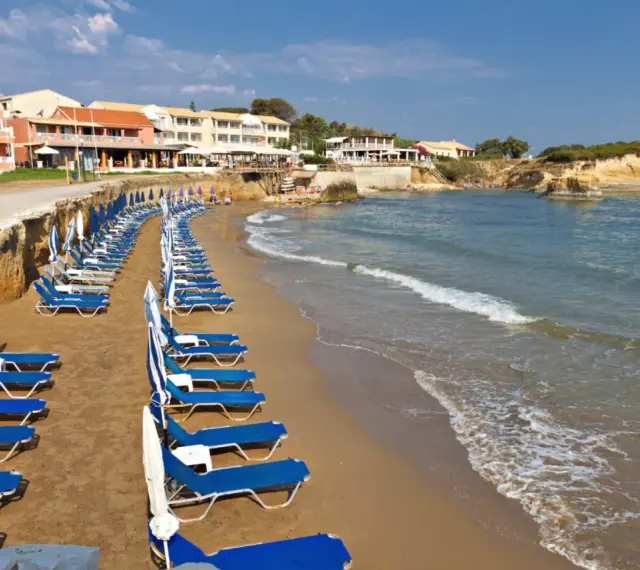 Rzędy leżaków i parasoli na piaszczystej plaży, z hotelami w tle. Idealne miejsce, by odpocząć i zastanowić się, gdzie na Korfu wybrać się na wakacje.