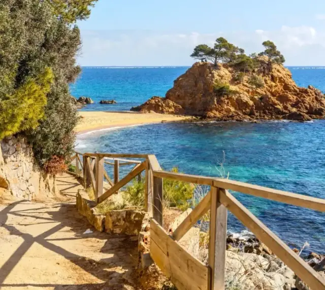 Ścieżka z drewnianą balustradą prowadzi do urokliwej zatoczki z piaszczystą plażą i skalistą wysepką. To idealne miejsce na odkrywanie uroków Costa Brava.