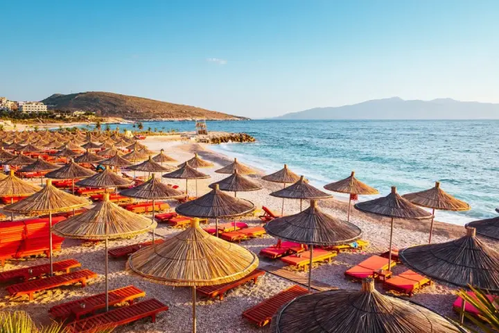 Albania, lipiec: słoneczna plaża z rzędami parasoli i leżaków, turkusowe morze i góry w tle. Idealna pogoda na wakacje.