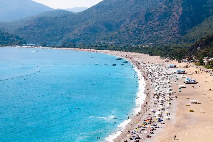 Plaża z turkusową wodą, górami w tle i tłumem ludzi odpoczywających pod parasolami.
