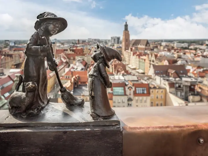 Figurki krasnali: czarownica z kotem i miotłą, na tle panoramy Wrocławia.