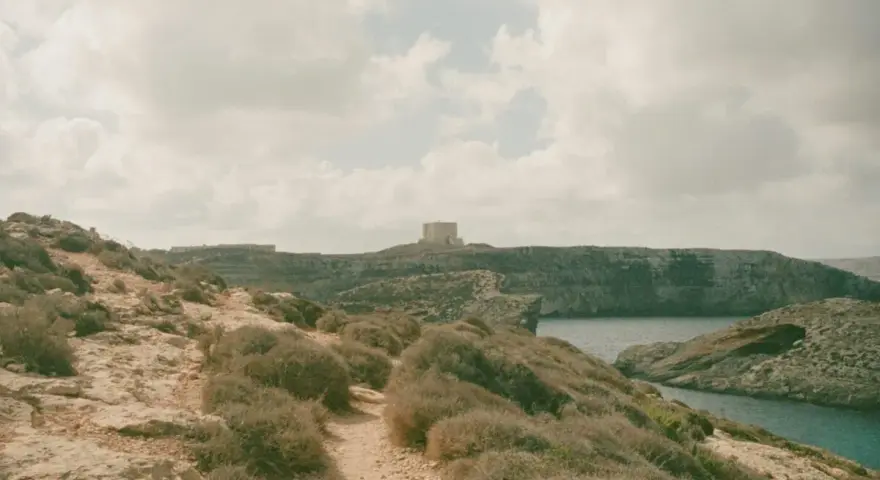 Malta czy warto? Kamienista ścieżka prowadzi przez skaliste wybrzeże z niską roślinnością do zatoki. W oddali widać wieżę obronną.