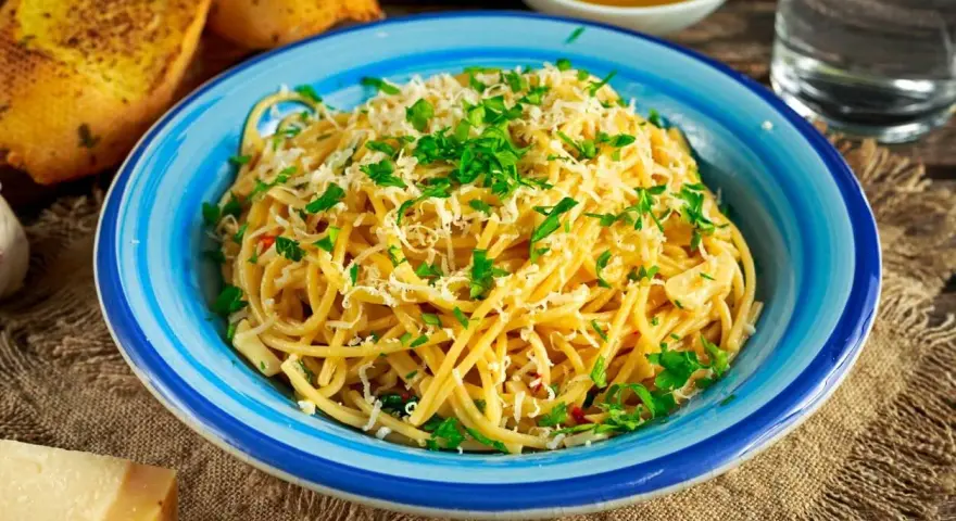 Pyszne spaghetti aglio olio posypane parmezanem i natką pietruszki, podane w niebieskiej misce. Obok grzanki czosnkowe i kawałek sera.