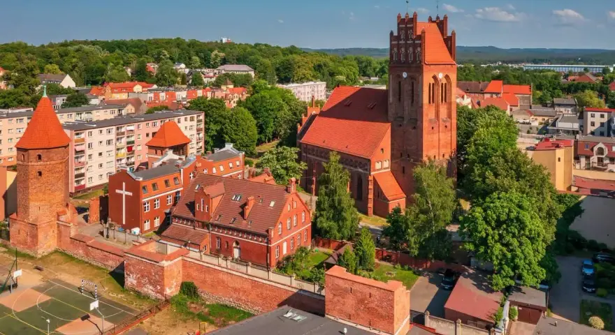 Zamek krzyżacki w Lęborku, z charakterystyczną wieżą i ceglaną architekturą, otoczony zielenią i zabudowaniami miejskimi.