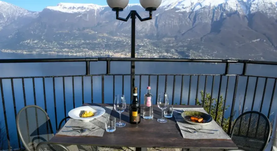 Stół z winem i jedzeniem na tarasie z widokiem na jezioro i ośnieżone góry. Idealne miejsce na romantyczną kolację z widokiem, prawdziwa "terrazza del brivido".