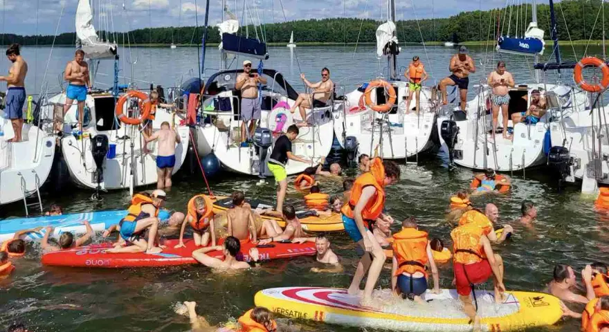Ludzie w kamizelkach ratunkowych i strojach kąpielowych pływają i bawią się na wodzie przy żaglówkach.