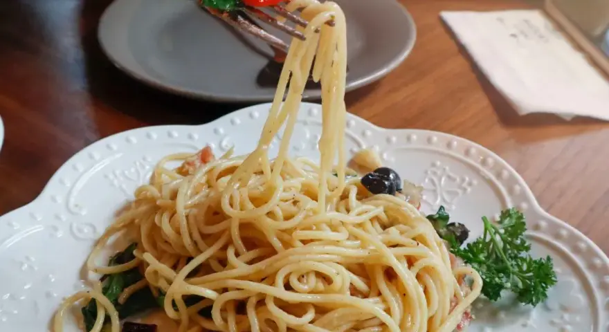 Widok z bliska na talerz z makaronem aglio olio, z widelcem unoszącym porcję spaghetti z bazylią i pomidorkiem.