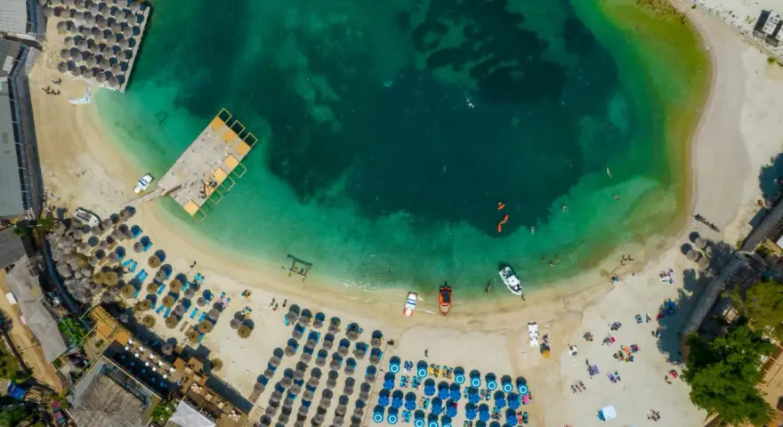 Widok z lotu ptaka na zatokę z turkusową wodą, plażą z leżakami i parasolami oraz łodziami.