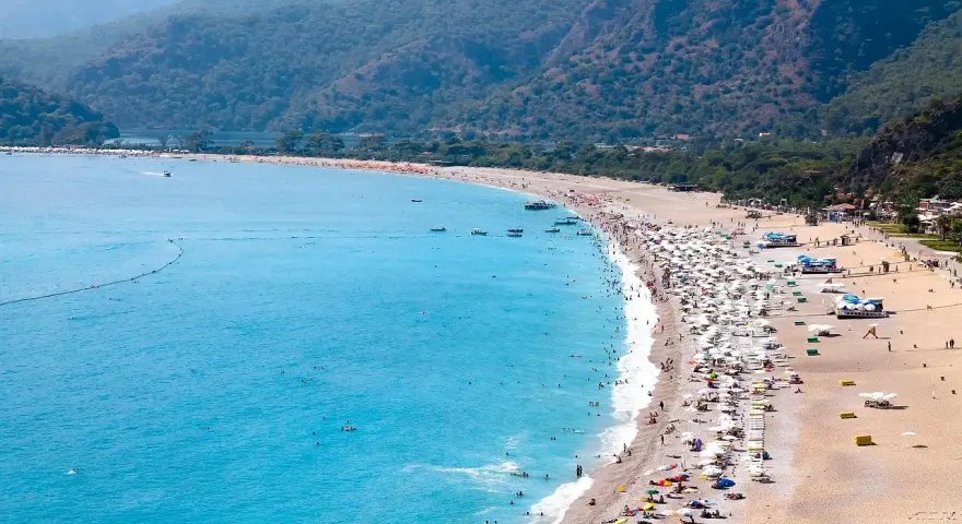 Plaża z turkusową wodą, górami w tle i tłumem ludzi odpoczywających pod parasolami.