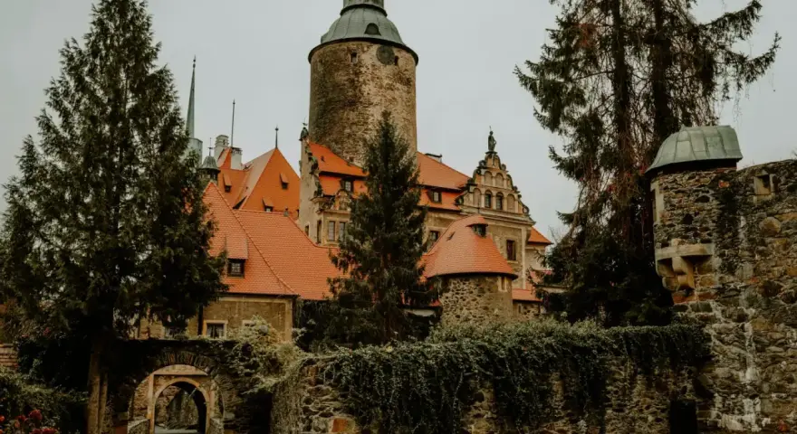 Kamienny zamek z wieżą i czerwonymi dachami, otoczony drzewami i murem.